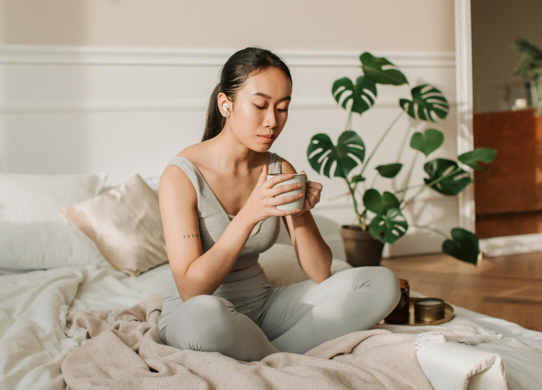 Woman sitting on a bed, holding a cup of tea. The room looks very calm and serene.