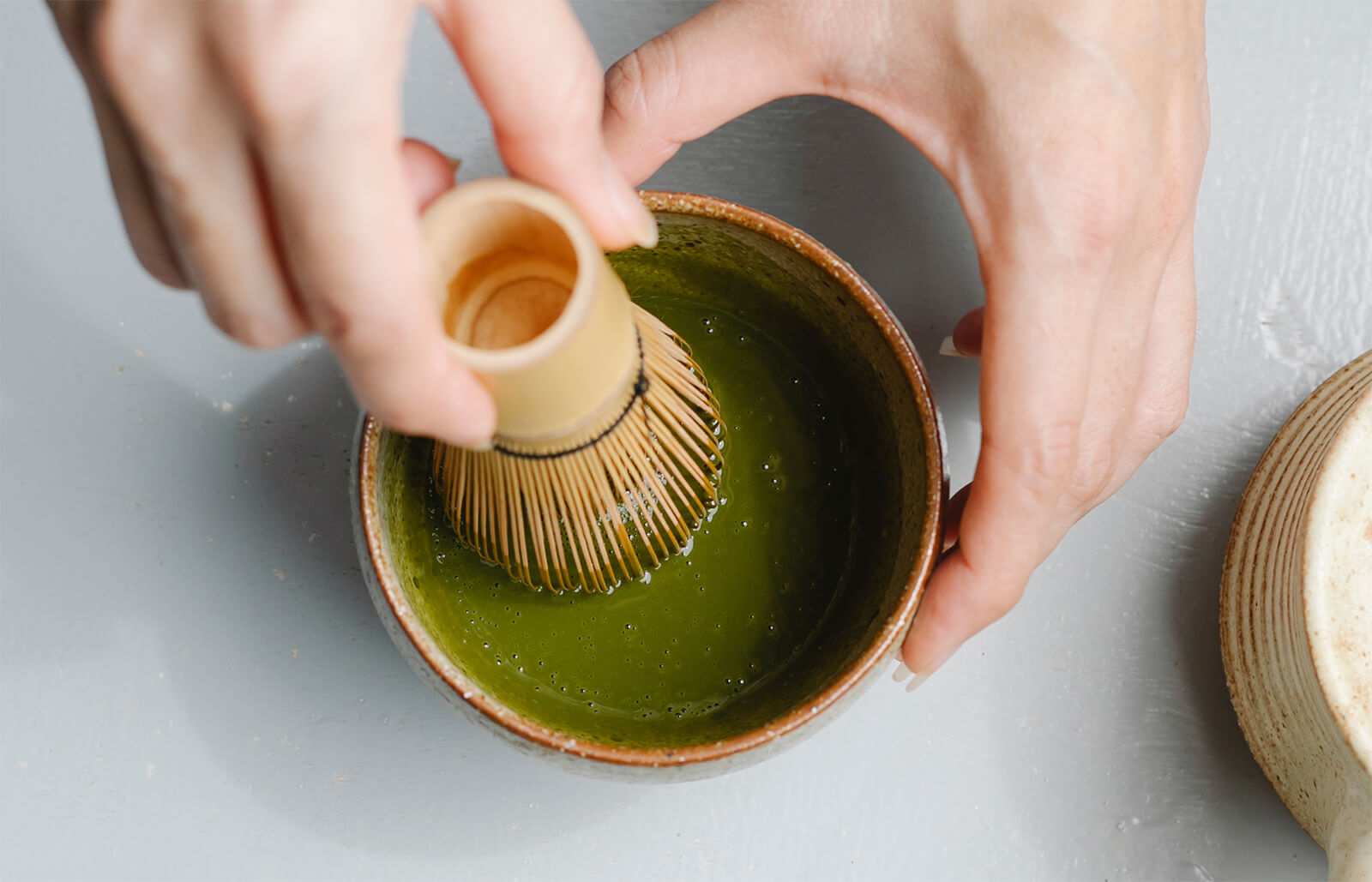 an image of a person’s hands holding a cup of matcha, and whisking it with traditional bamboo whisk.