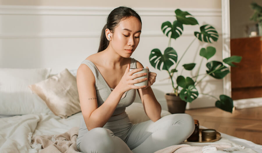 Woman sitting on a bed, holding a cup of tea. The room looks very calm and serene.