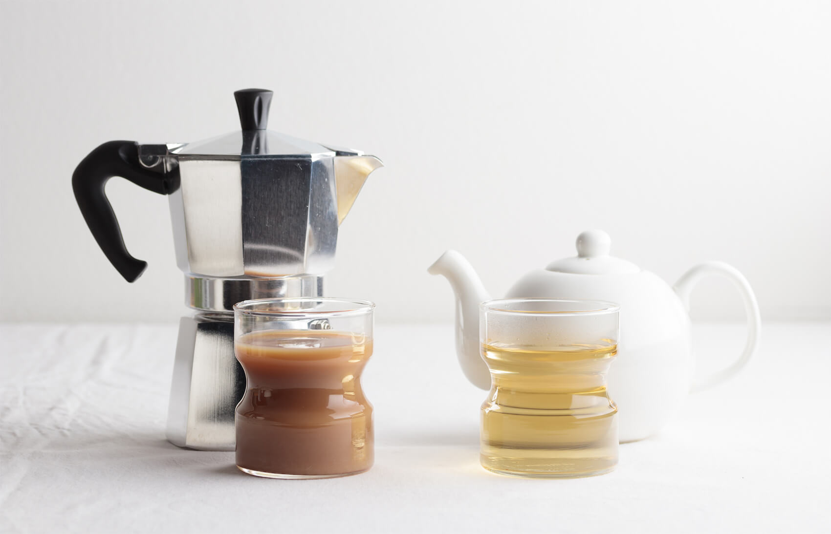 A photo of a coffee maker and tea pot. There are also 2 glasses: one filled with coffee, one with green tea.